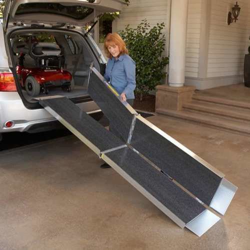 A woman stands beside an open SUV trunk in a driveway, using a foldable metal and carpeted vehicle ramp to access the back. A red wheelchair is visible inside the vehicle.
