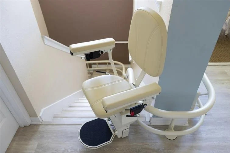 A cream-colored stair lift chair mounted on a staircase with a handrail, shown in an indoor residential setting with light wood flooring and beige walls.