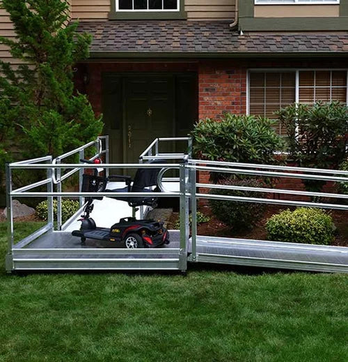A metal wheelchair lift ramp installed in front of a residential house, shown with a wheelchair being transported up the ramp onto a porch.