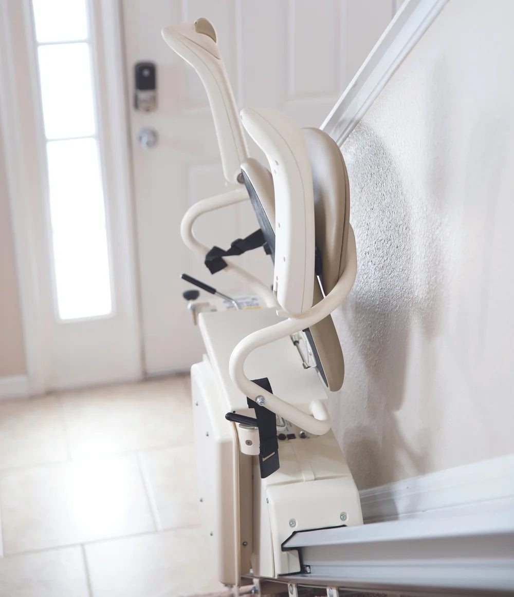 A cream-colored indoor staircase lift mounted on a wall beside a staircase, shown in a residential hallway with a door and tiled floor in the background.