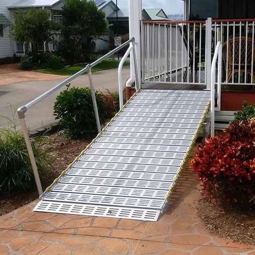 A metal utility ramp with a slotted grid design, installed on a stone patio leading up to a house porch with white railings and landscaping.