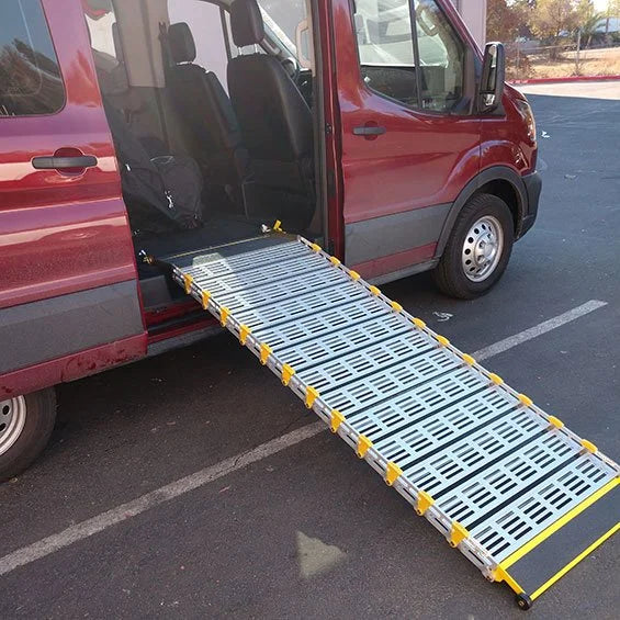 A metal vehicle ramp with yellow safety markings is deployed from the open side door of a red van parked on an asphalt lot.
