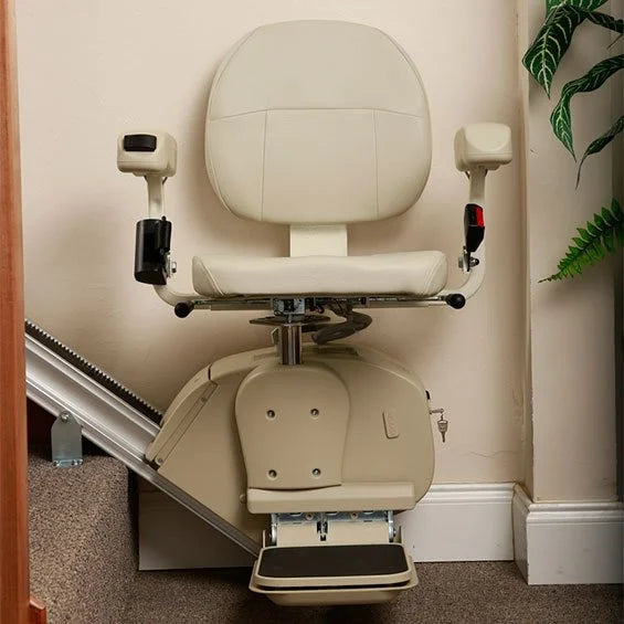 An indoor stairlift chair with a cream-colored upholstered seat and backrest, mounted on a metal stair rail against a beige wall with carpeted flooring and a plant visible at the side.