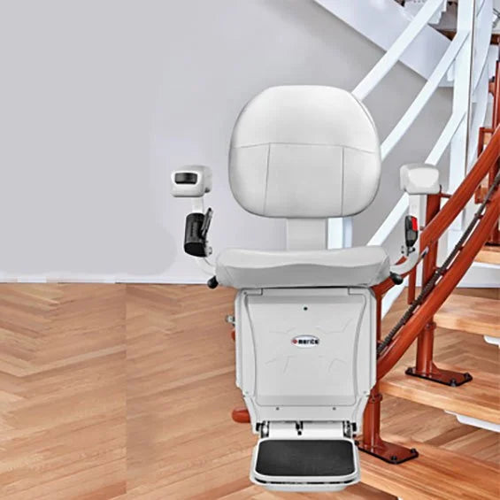 A white stair chair lift with a padded seat and backrest, mounted beside wooden stairs with a white railing in an indoor setting.